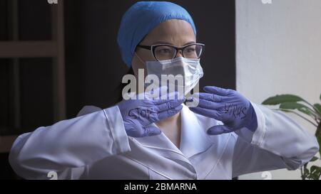 Le médecin féminin fatigué en uniforme blanc et masque protecteur montre l'inscription virus stop sur ses paumes dans des gants en caoutchouc regardant sur le côté Banque D'Images