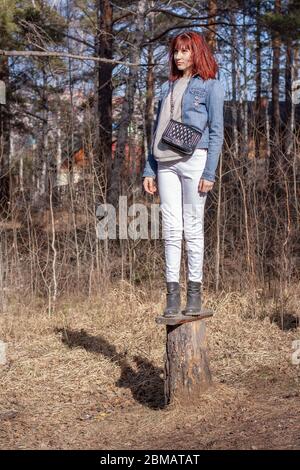 Une jeune fille à cheveux rouges se tient sur une souche dans la forêt. Apparence européenne. Veste en denim et pantalon blanc. Sac à main sur l'épaule. Verticale. Banque D'Images