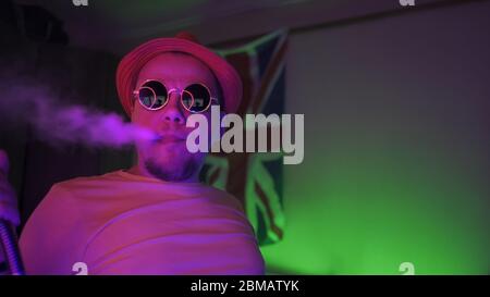 Fumeur de Hookah exhalant fumée dans Neon Lighting. Homme dans des lunettes de soleil en auto-isolation à la maison fume dans la chambre avec des lumières néon sur le drapeau GB arrière-plan Banque D'Images