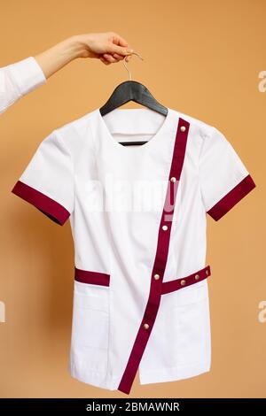 La main féminine tient une cintre avec une chemise blanche à bordure bordeaux pour les travailleurs médicaux sur fond orange. Banque D'Images