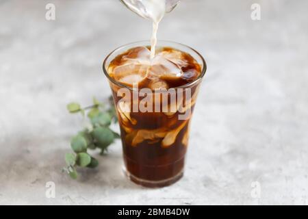 Le lait est versé dans un grand verre avec du café noir préparé avec des morceaux de glace sur fond gris foncé, gros plan. Concept de café frais d'été glacé Banque D'Images