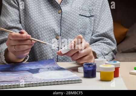 Une fille en pyjama à une table dessine un gros plan tenant un pinceau et un chapeau d'un pot d'aquarelle dans ses mains et mélange les peintures dans les tons bleus. Dur. Occupation Banque D'Images