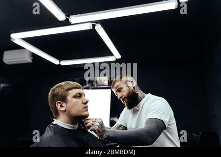 Barbu tatttooed Barber taming homme avec machine à raser dans le barbershop. Processus de coiffure. Stylist coupe la barbe d'un homme barbu. Banque D'Images