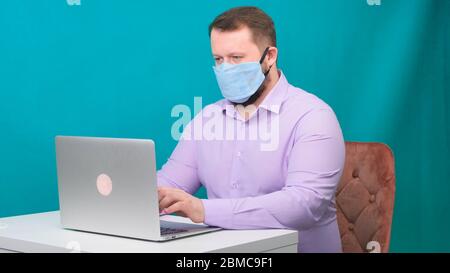 Portrait d'un homme de brunette gai avec masque médical chirurgical travaillant sur la saisie d'un ordinateur portable de messagerie ou de discuter en social. Banque D'Images