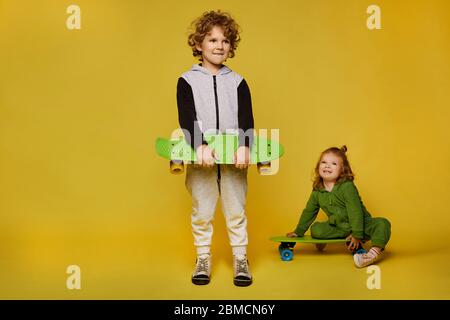 Enfants élégants en costume modish posant avec des planches à roulettes. Petite fille et petit garçon isolés sur fond jaune avec espace de copie. Enfants Banque D'Images