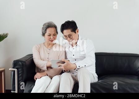 Un couple asiatique âgé utilisant des appareils électroniques sur le canapé Banque D'Images