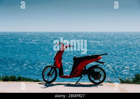 Scooter rouge stationné sur une plage verte herbacée à côté de l'océan, par une journée d'été ensoleillée avec un ciel bleu Banque D'Images