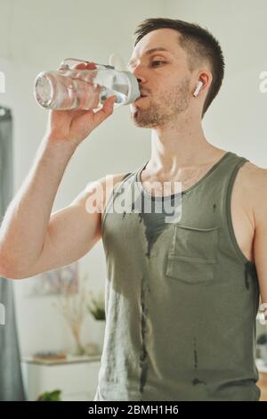 Jeune sportif sueur dans des écouteurs et des vêtements actifs eau potable de bouteille après un entraînement dur à la maison pendant la période de quarantaine Banque D'Images