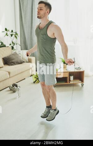 Jeune athlète contemporain en short, gilet de sport et baskets sautant avec corde à sauter dans le salon pendant l'entraînement le matin Banque D'Images