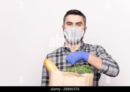 Un jeune messager masculin dans un masque de protection et des gants livrant du pain et des légumes frais à votre maison dans un sachet de papier isolé Banque D'Images