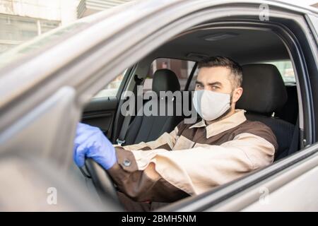 Jeune homme en gants de protection, masque et vêtements de travail vous regardant en étant assis en voiture et en conduisant à des clients pour leur livrer des commandes en ligne Banque D'Images