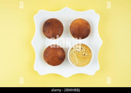 Trois muffins faits maison et une tasse de café sur une assiette carrée blanche sur fond jaune. Banque D'Images