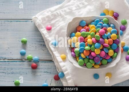 Vue de dessus d'un bol rempli de bonbons colorés sur un tapis blanc et de bois vintage avec quelques uns éparpillés autour. Banque D'Images
