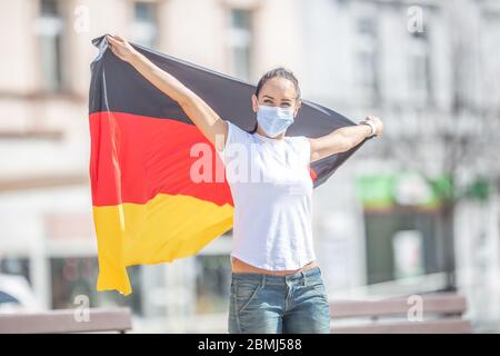 Fan allemand dans un masque de visage tient le drapeau derrière elle dehors, souriant. Banque D'Images