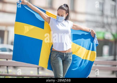 Brunetter debout à l'extérieur regarde le drapeau suédois qu'elle tient, avec un masque facial. Banque D'Images