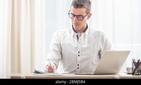 L'homme écrit dans son bloc-notes avec un carnet ouvert sur son bureau en face de lui. Banque D'Images