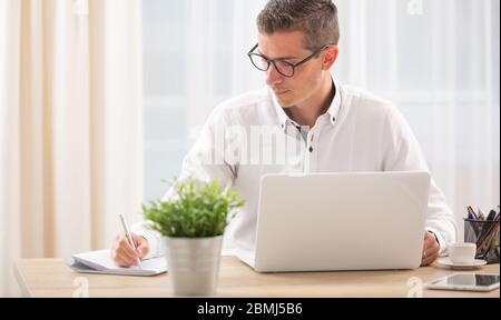 Homme écrit dans son bloc-notes avec un carnet ouvert sur son bureau en face de lui. Banque D'Images