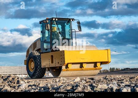 Un rouleau de route jaune accrocheur avec cabine climatisée fermée se dresse sur une route non prête, des pierres, un ciel bleu, des nuages, vue latérale avant gauche. C Banque D'Images
