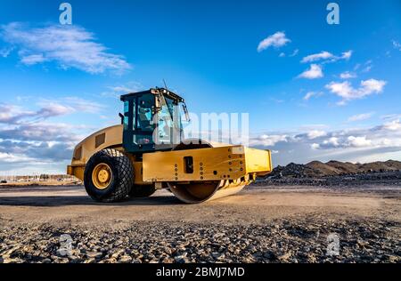 Un rouleau de route jaune accrocheur avec cabine climatisée fermée se dresse sur une route non prête, des pierres, un ciel bleu, des nuages, une vue latérale gauche. Nettoyer s Banque D'Images