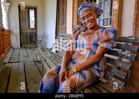 Portrait d'une femme d'âge moyen souriante vêtue d'une robe ethnique colorée, assise aux portes de sa maison coloniale de Creektown. Banque D'Images
