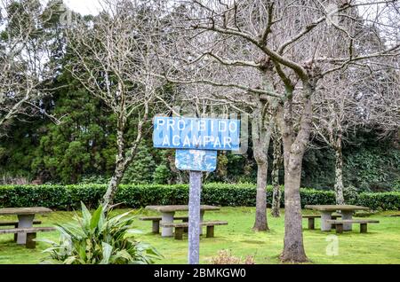 Vieux signe bleu avec texte Proibido Acampar en langue portugaise. TRADUCTION: Camping interdit. Parc avec arbres verts, bancs et tables en arrière-plan. Pas de panneau de camping, pas de séjour de nuit. Banque D'Images