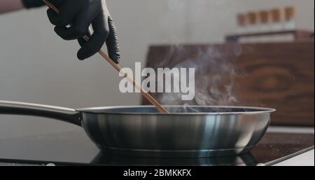 homme main dans des gants en déplaçant quelque chose avec une spatule en bois dedans poêle à frire Banque D'Images