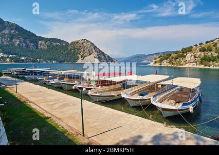 Petits bateaux amarrés dans un lagon bleu de mer Égée avec des îles montagneuses autour, plage en arrière-plan et drapeau turc en haut de la colline à Turunc, Mugla, Tu Banque D'Images