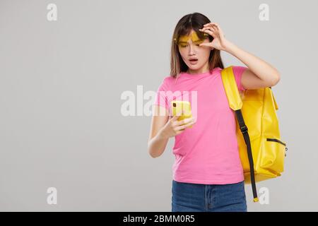 Une adolescente asiatique en lunettes en forme de cœur tient un téléphone portable dans les mains sur fond gris. Nouvelle publicité gadget Banque D'Images