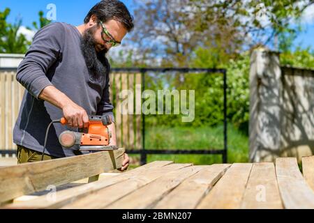 Homme de race blanche, adulte moyen, dans le jardin, ponçant des planches en bois. Bricolage rénovation, restauration, menuiserie concept. Banque D'Images