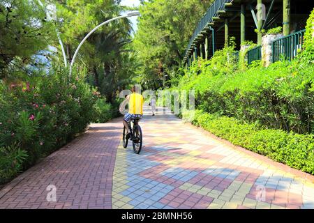 Un adolescent fait un vélo le long d'une piste cyclable dans un parc d'une ville de villégiature parmi les arbres. Vacances d'été Turquie, Marmaris. Banque D'Images