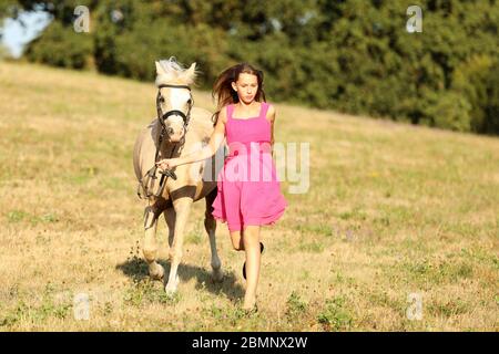 Jeune fille en robe rose courir avec poney à travers le pré dans l'après-midi ensoleillé d'été Banque D'Images