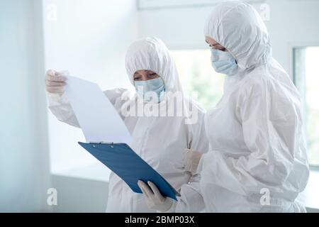 Travailleurs médicaux en vêtements de protection et masques médicaux regardant à travers les papiers sur la chemise de clip Banque D'Images