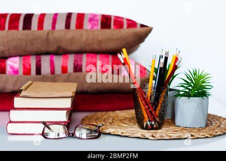 Décoration intérieure chaleureuse : crayons dans le support, pile de livres, plantes en pots sur un support en osier, oreillers et écossais sur une table blanche. Distance home éduca Banque D'Images