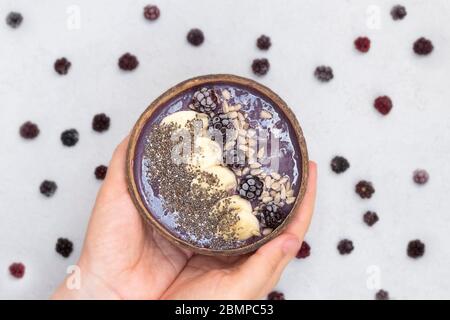 Femme mains tenant un bol de noix de coco avec smoothie de mûre avec banane surgelée, mûres et graines, vue du dessus Banque D'Images