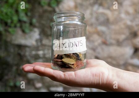 Un pot en verre avec des pièces de monnaie à l'intérieur et une étiquette de vacances sur une main, sur une plante et un fond de mur de pierre. Un bote de cristal con monedas dentro, tiene una et Banque D'Images