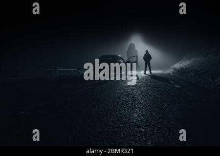 Une femme fantôme floue dans une robe debout flottant sur une route de campagne la nuit. Avec une voiture et un conducteur derrière. Banque D'Images