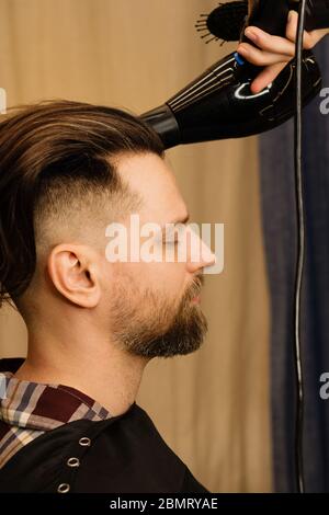 Beau barbu homme tout en ayant ses cheveux coupés par le coiffeur au salon de coiffure. Vue rapprochée de l'homme barbu en train de se faire coiffer avec sèche-cheveux au salon de coiffure. Banque D'Images