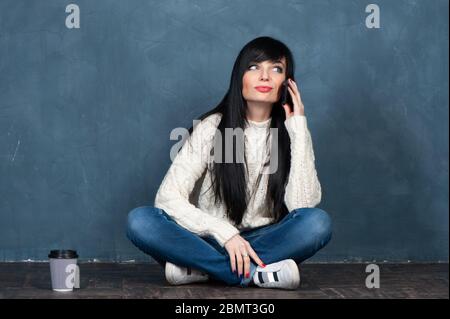 Une jeune fille brune tendance est assise sur le sol, parle au téléphone et sourit. Portrait de studio sur un fond bleu monochrome, branché et un espace pour le texte Banque D'Images