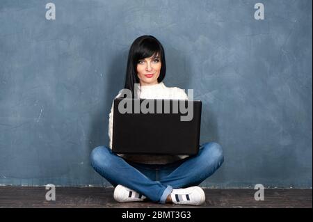 Belle fille brune est assise sur le sol avec son ordinateur portable, regarde dans l'appareil photo et sourit. Portrait de studio sur un noir monochrome, shabby backgro tendance Banque D'Images