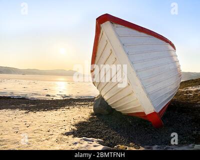 Photo d'un bateau en bois sur le bord de mer de Magadan, Russie. Bateau blanc à rayures rouges. Coucher de soleil sur l'arrière-plan. Ciel bleu clair et glace sur la mer en hiver Banque D'Images
