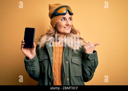 Femme de ski blonde d'âge moyen portant des lunettes de ski tenant un smartphone montrant l'écran pointant et montrant avec le pouce jusqu'au côté avec Happy face smi Banque D'Images