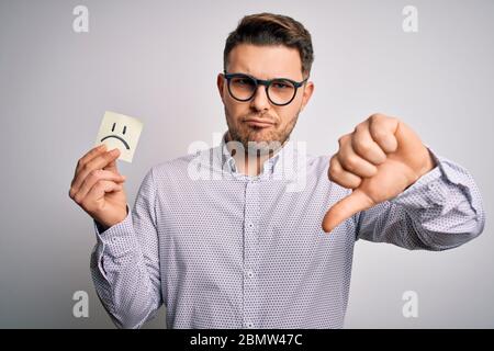 Jeune homme d'affaires avec les yeux bleus tenant une note en papier avec le visage triste comme message malheureux avec le visage en colère, signe négatif montrant le mécontentement avec les pouces vers le bas, Banque D'Images