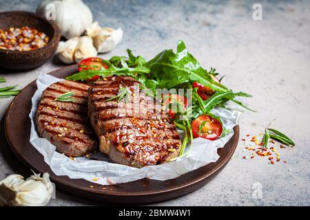 Steaks de bœuf grillés avec des roquette et salade de tomates sur une assiette en bois, fond sombre. Banque D'Images