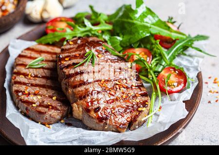 Steaks de bœuf grillés avec des roquette et salade de tomates sur une assiette en bois, fond sombre. Banque D'Images