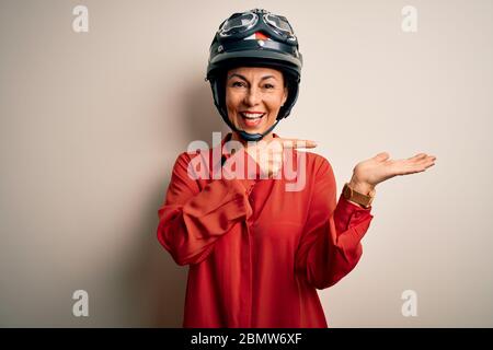 Femme motard d'âge moyen portant un casque de moto sur un arrière-plan blanc isolé stupéfait et souriant à la caméra tout en présentant avec la main et Banque D'Images