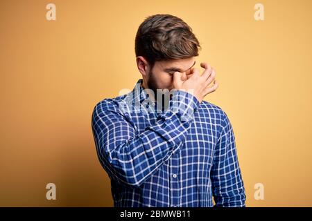 Jeune homme d'affaires blond avec barbe et yeux bleus portant une chemise sur fond jaune fatigué frottant nez et yeux sensation de fatigue et de maux de tête. Stress Banque D'Images