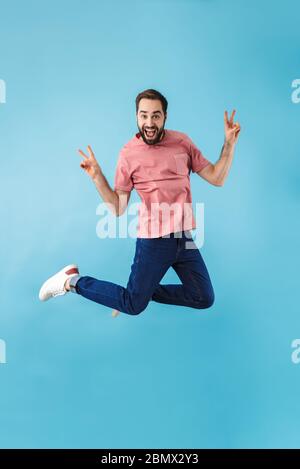 Image d'un jeune beau heureux heureux excité homme sautant isolé sur fond bleu mur faire le geste de paix. Banque D'Images