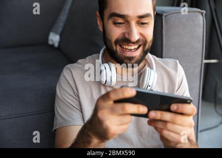 Photo d'un jeune homme optimiste heureux à l'intérieur à la maison jeux de jeu par téléphone portable assis sur le sol. Banque D'Images