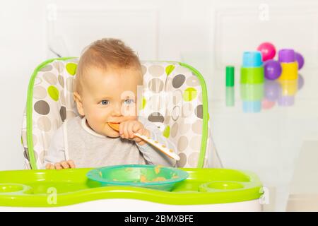 Un tout-petit garçon ayant un aspect réfléchi et une cuillère en plastique pour bébé dans la bouche s'assoient dans une chaise haute avec une assiette de porridge Banque D'Images