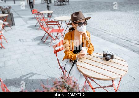 Jeune femme au masque facial, assise sur la terrasse du café seule. Concept de distanciation sociale et nouvelles règles sociales après la pandémie du coronavirus. Banque D'Images
